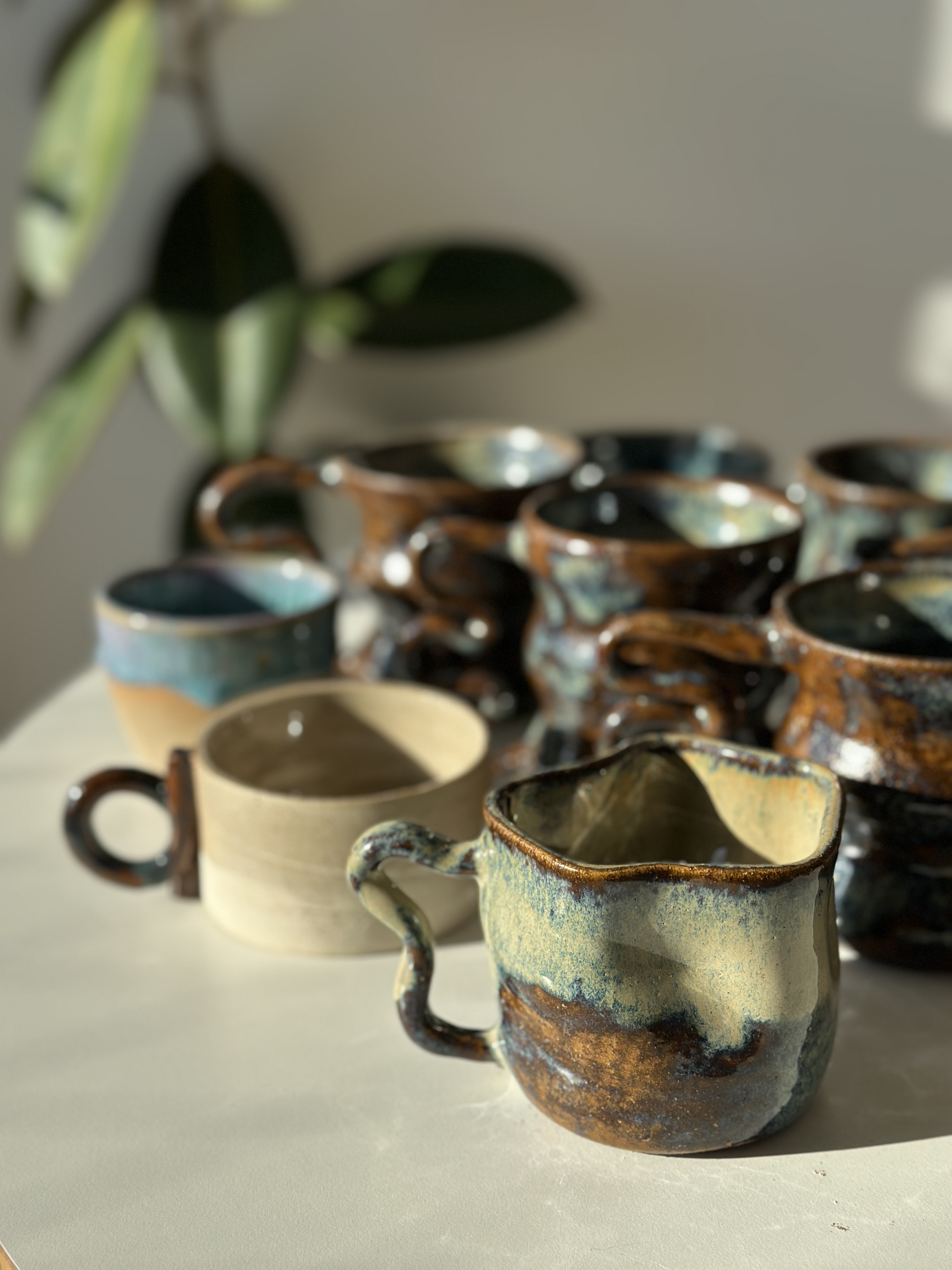 Closing image · finished Käffi mugs and a Flux pour-vessel resting in warm side light, plant in soft focus behind.