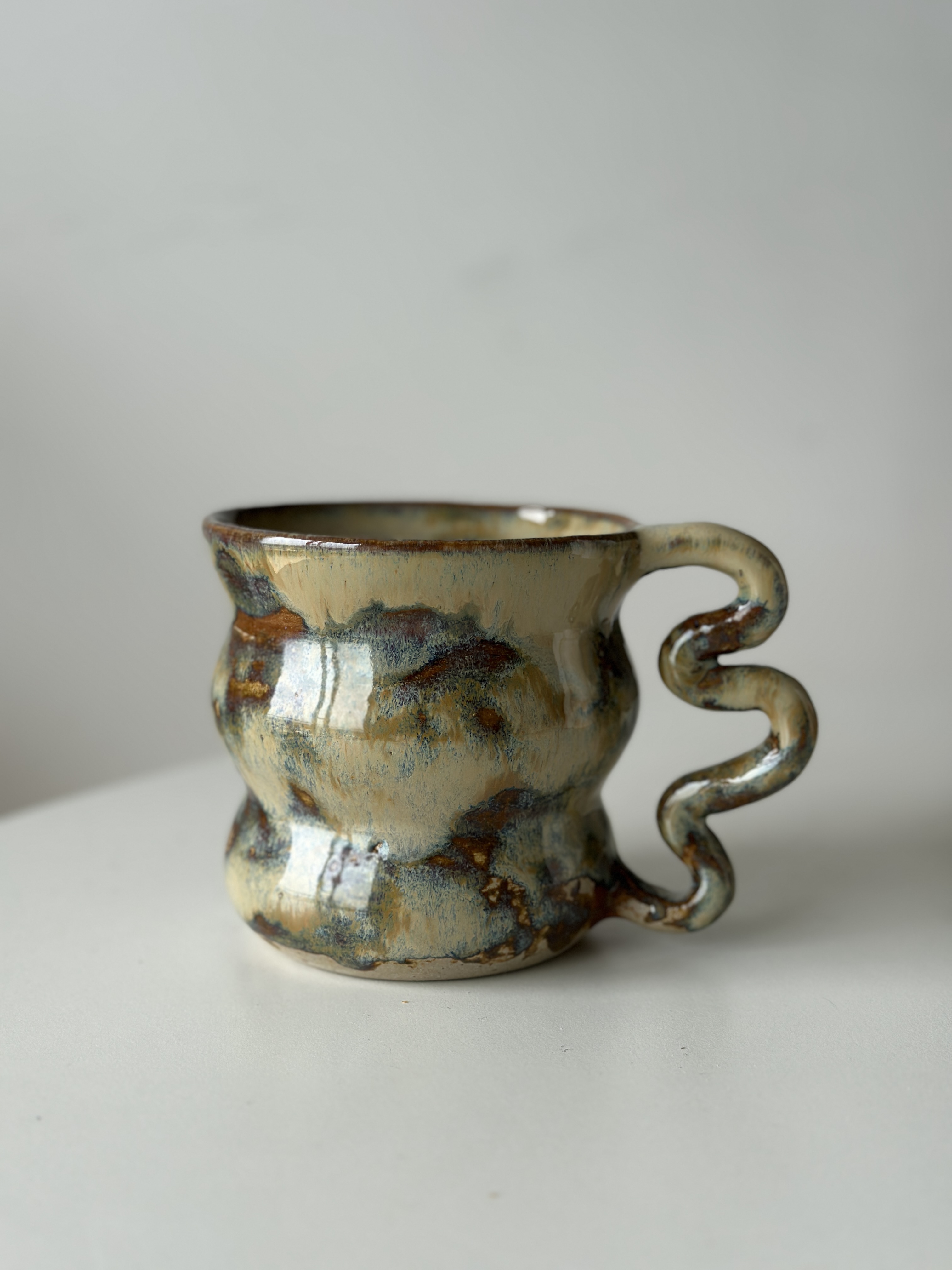 Greytà ceramic mug with wavy curling handle, iridescent earth-tone glaze, on a soft neutral background.
