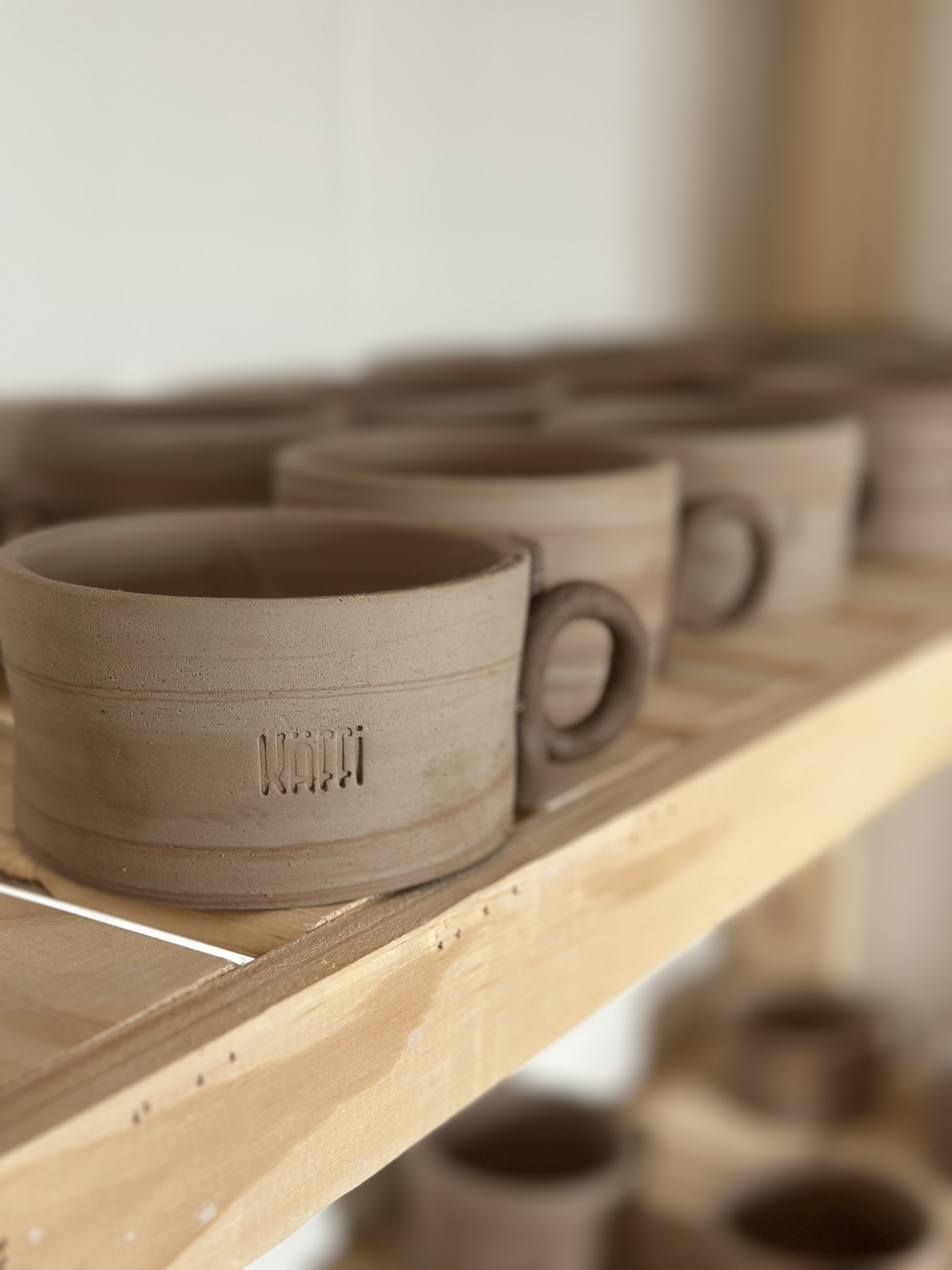 Käffi greenware · stoneware mugs lined up on a wood drying rack with engraved Käffi mark.