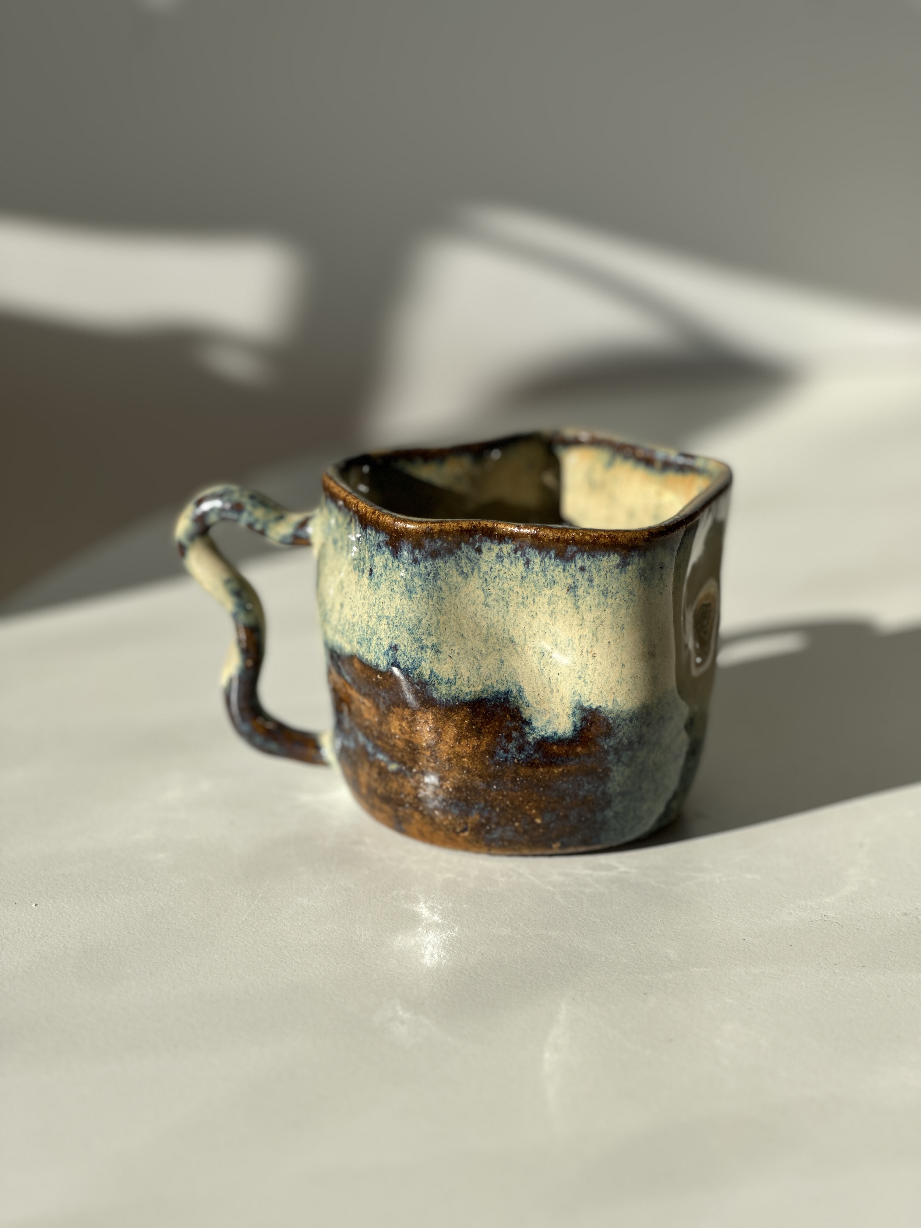 Taza Eco · stoneware mug with copper and pale aqua drip glaze and twisted vine handle, in warm side light.