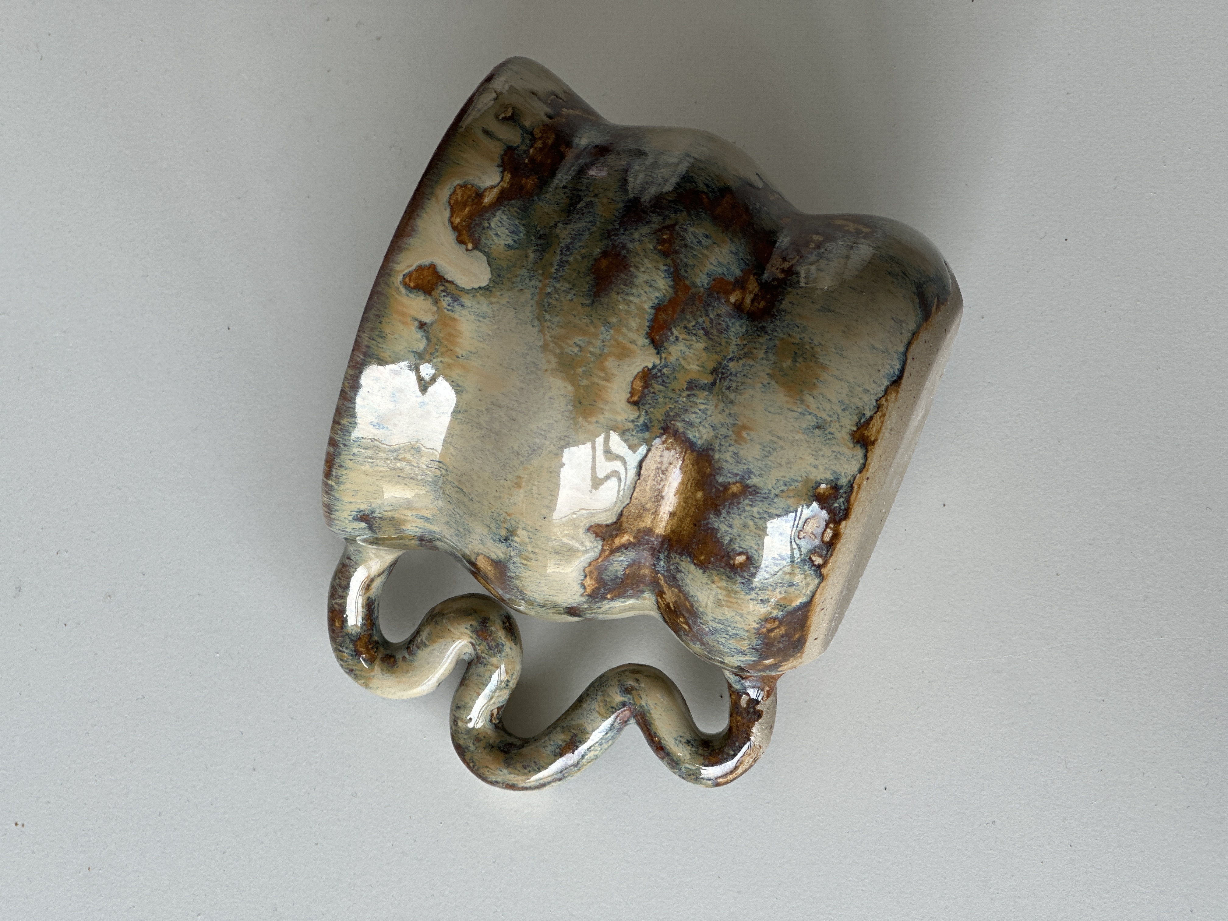 Taza Flux · iridescent earth-tone glaze with wavy curling handle, top-down view.